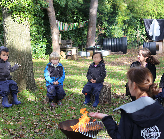 Children and adults sitting on logs are gathered around a fire pit in a natural outdoor setting.