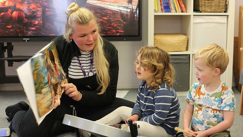 
An adult is reading a storybook to two young children in a classroom setting.
