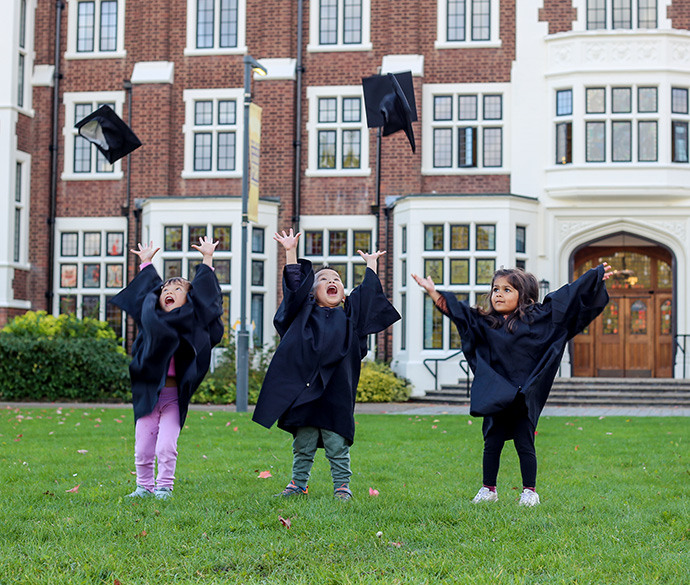 Three children standing on a lawn in graduation gowns tossing their caps in the air. There's a large building with many windows in the background.