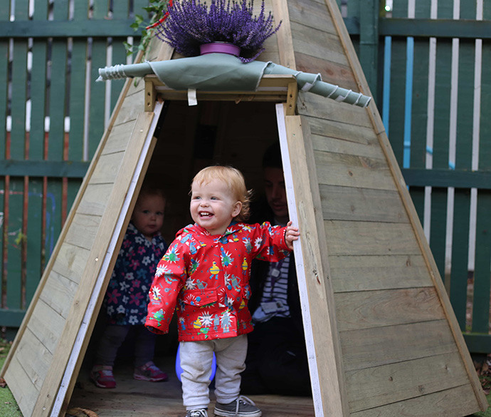A child with a big smile wearing a colourful coat stands at the entrance of a wooden playhouse, while another child and an adult are visible inside the playhouse behind them.