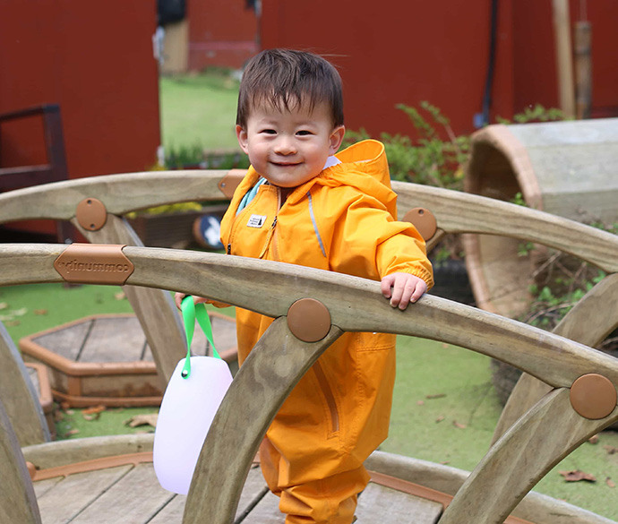 A smiling child in an orange rainsuit standing on a wooden playground bridge, holding a white lantern with a green handle.