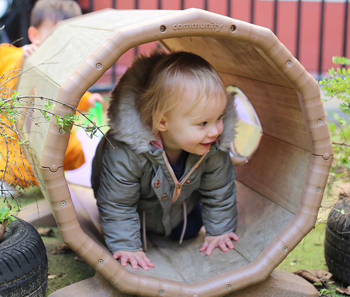 A young child wearing a coat is crawling through a tunnel-like structure in an outdoor playground.