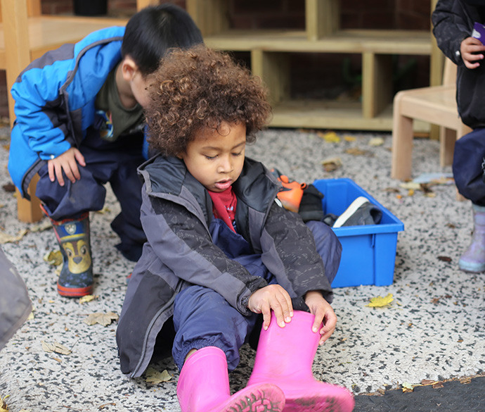 A child in a coat is sitting down putting on pink wellington boots. There are two children wearing coats and wellington boots in the background, one is reaching for an item in a blue box.