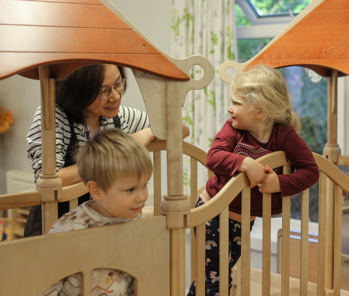 An adult is smiling and interacting with two children on a wooden indoor playground structure.