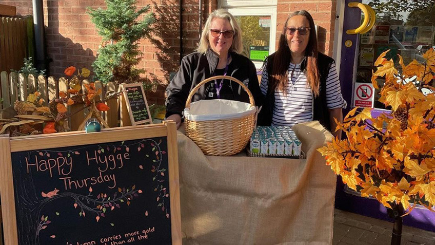 
Two smiling people standing behind an outdoor display featuring a wicker basket, autumn foliage, and a chalkboard reading "Happy Hygge Thursday".
