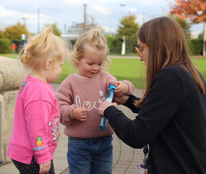 
A person with glasses kneels outdoors, helping two young children open a small blue bubble wand container. Grass and trees are in the background.
