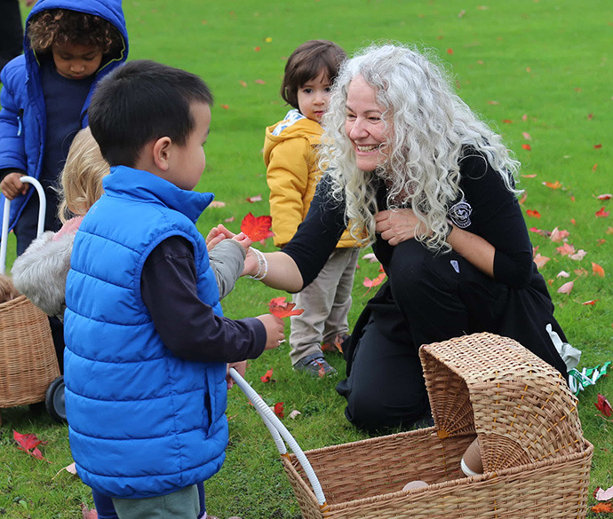 
A person kneels on grass, smiling and greeting a young child in a blue vest. Other children and wicker baskets are nearby.
