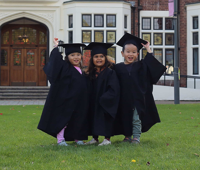 
Three young children in black graduation caps and gowns stand together smiling on a lawn in front of a large building with many windows.
