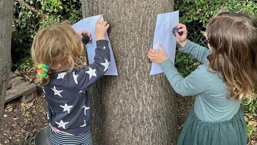 
Two children are each holding a piece of paper and a crayon over a tree trunk for nature rubbings.
